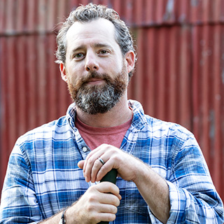 A white man with a graying beard wearing a flannel shirt stands in front of a red barn.